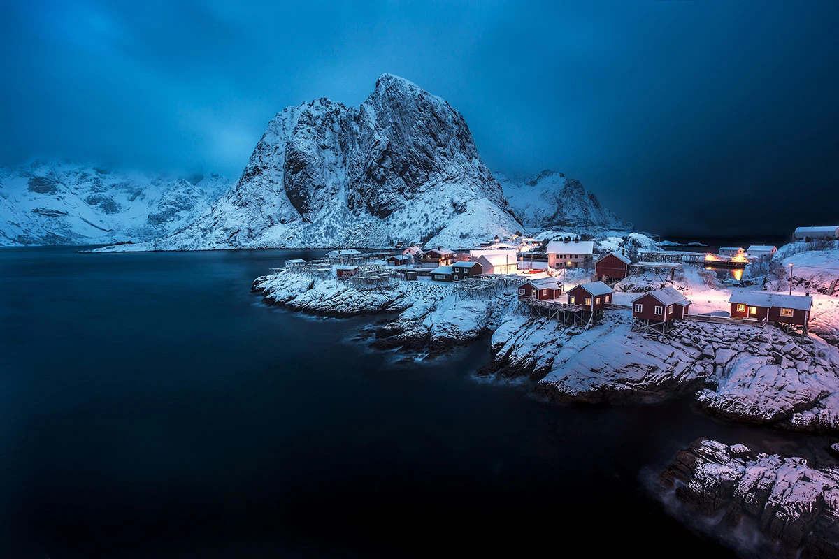 Snødekt kystlandskap i blått kveldslys med et lite fiskevær av røde rorbuer langs svabergene, varmt lys i vinduene og høye, dramatiske fjell som reiser seg bak den rolige fjorden.